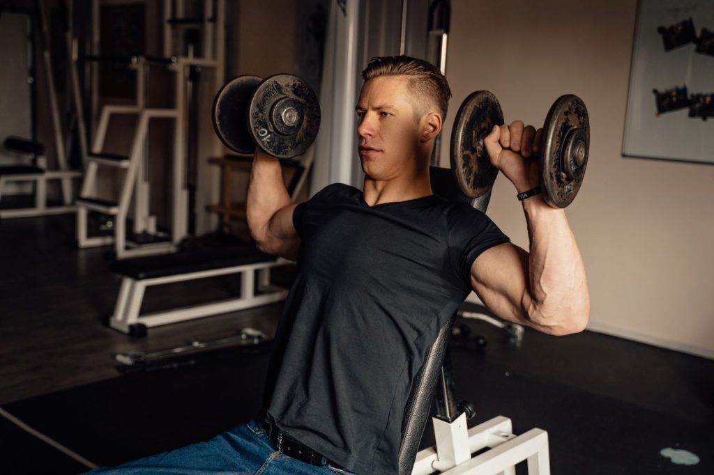 A focused man in a gym performing a dumbbell shoulder press, showcasing fitness and strength.